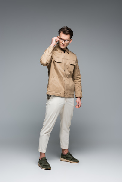 повна довжина модного чоловіка в стильному вбранні позує під час регулювання окулярів на сірому
 - Фото, зображення
