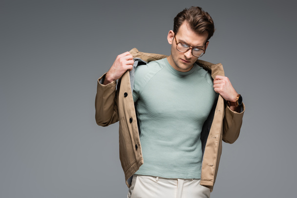 trendy man in glasses adjusting jacket while posing isolated on grey - 写真・画像