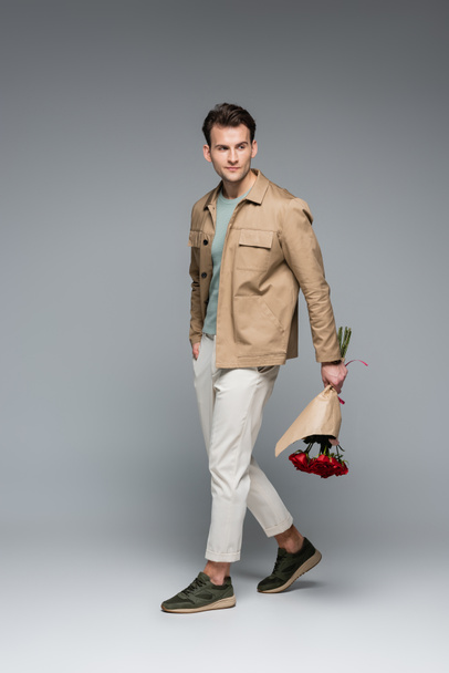 full length of trendy man holding bouquet of red roses wrapped in paper while walking on grey - Foto, imagen