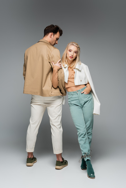 full length of stylish model in pants and jacket leaning on man while posing on grey - Zdjęcie, obraz