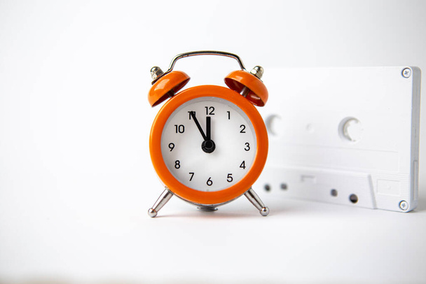 Velho relógio laranja analógico sobre um fundo branco. O relógio mostra o tempo de cinco minutos a doze horas, e ao lado dele é uma cassete de música de áudio branco. Espaço Copiar. Relógio. O tempo. Música. - Foto, Imagem