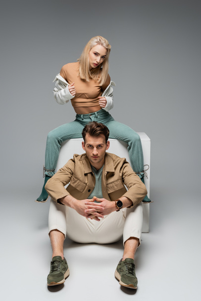 Полная длина стильный мужчина и женщина в брюках и куртках, позируя возле белого куба на сером - Фото, изображение