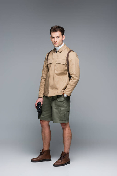 full length of trendy traveler in shorts and jacket standing with hand in pocket while holding binoculars on grey  - Foto, immagini