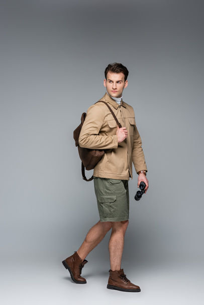 full length of trendy traveler in shorts and jacket walking with binoculars on grey  - Photo, Image