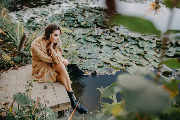 若い女性は水ユリと池で木製の桟橋に座ってマキシシフォンドレスを着て. - 写真・画像