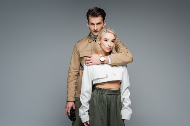 trendy man holding binoculars and hugging blonde woman isolated on grey  - Fotografie, Obrázek