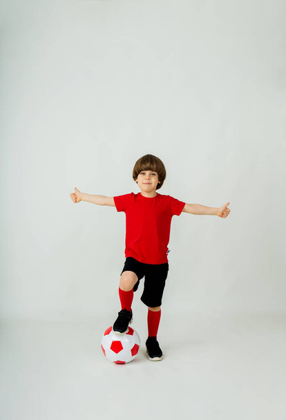 Kırmızı tişörtlü ve şortlu mutlu bir futbolcu beyaz arka planda futbol topuyla duruyor. - Fotoğraf, Görsel