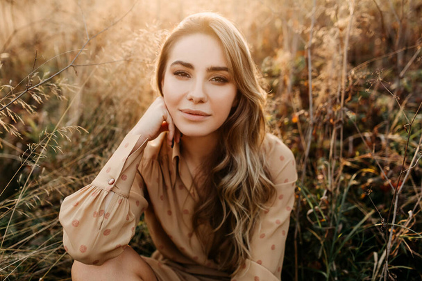 Young woman with long blonde wavy hair, smiling, sitting in a field with tall dry grass. - Foto, Imagem