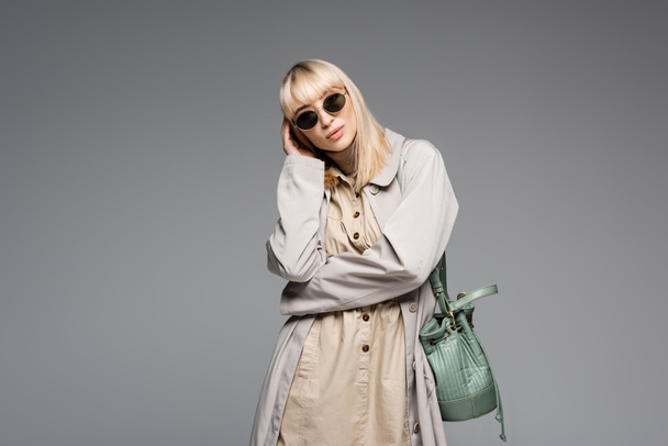fashionable woman in sunglasses and trench coat posing with green bag while standing isolated on grey - 写真・画像