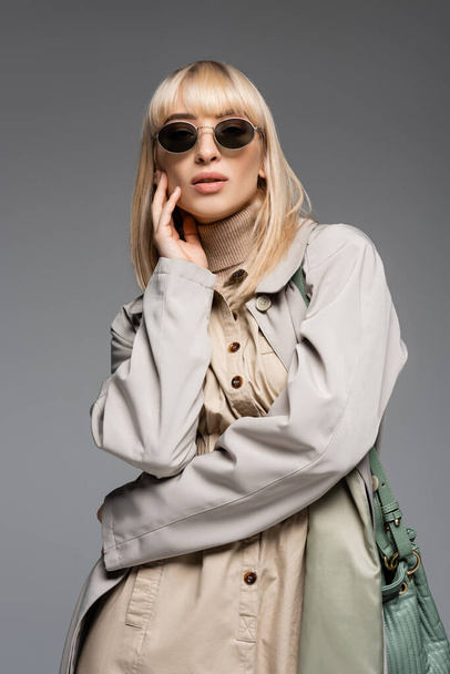 young fashionable woman in sunglasses and trench coat posing isolated on grey - 写真・画像