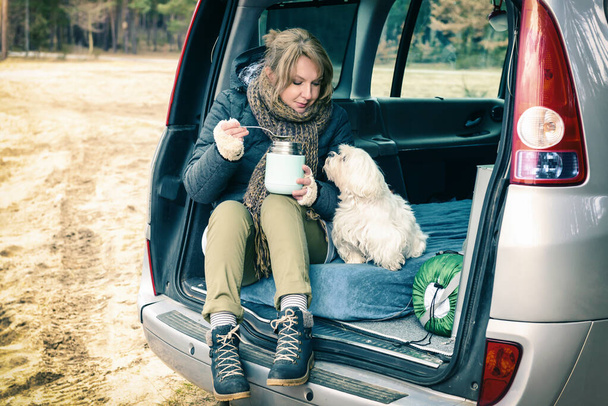 Vrouw die een warme schotel uit een thermoskan eet tijdens een roadtrip, zittend in de kleine camper met hond, bos op de achtergrond - Foto, afbeelding