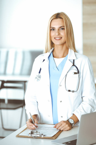 Doctor woman at work in hospital excited and happy of her profession. Blonde physician controls medication history records and exam results while using clipboard. Medicine and healthcare concept - Foto, Bild