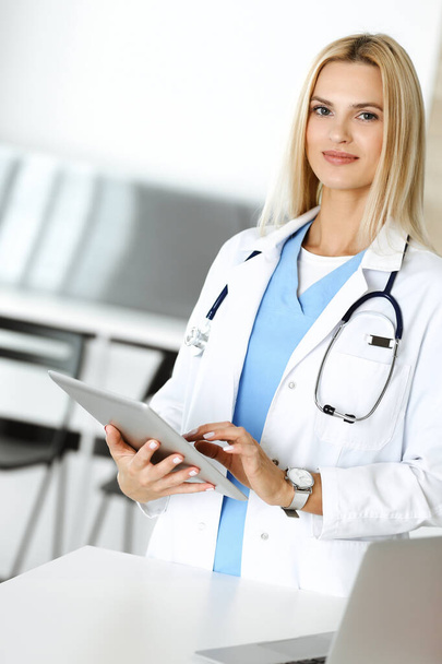 Woman-doctor at work in clinic excited and happy of her profession. Blond female physician is smiling while using tablet computer. Medicine concept - Foto, afbeelding