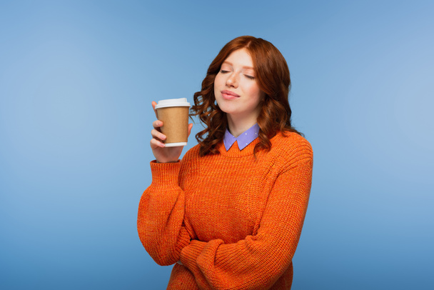pleased redhead woman in orange sweater holding coffee to go isolated on blue - Zdjęcie, obraz