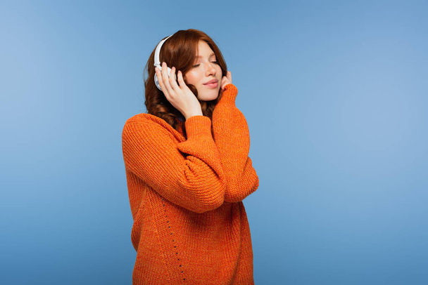 heureux rousse femme avec les yeux fermés écouter de la musique dans les écouteurs sans fil isolé sur bleu - Photo, image