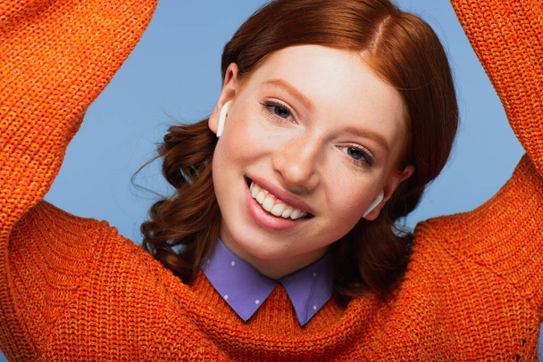 cheerful redhead woman listening music in wireless earphones isolated on blue - Fotó, kép