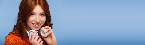 mulher ruiva positiva segurando fones de ouvido sem fio isolados em azul, banner - Foto, Imagem