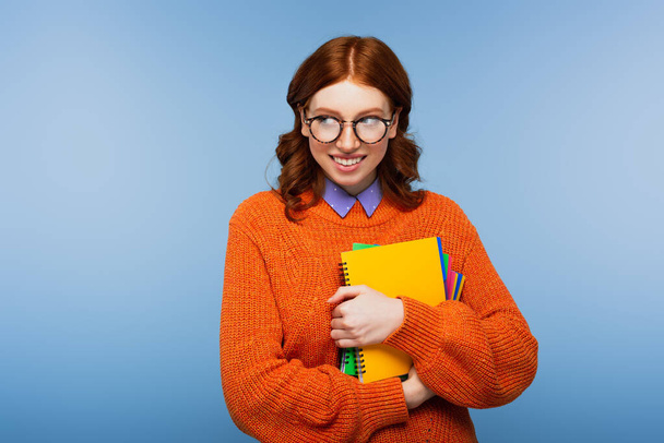 usmívající se zrzka student v brýlích a oranžový svetr drží notebooky a tužku izolované na modré - Fotografie, Obrázek