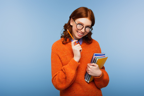 fröhliche rothaarige Studentin in Brille und orangefarbenem Pullover mit Notizbüchern und Bleistift auf blauem Hintergrund - Foto, Bild