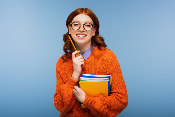 happy redhead student in glasses and orange sweater holding notebooks with pencil isolated on blue - Photo, Image