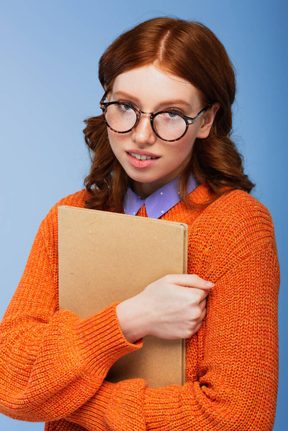 redhead student in glasses and orange sweater holding book isolated on blue - Photo, Image