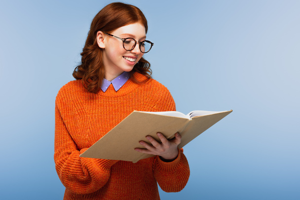 happy redhead student in glasses and orange sweater reading book isolated on blue - Фото, изображение