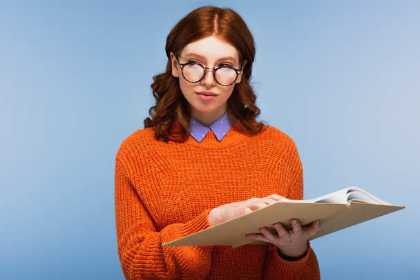 redhead student in glasses and orange sweater holding book and looking away isolated on blue - Photo, Image