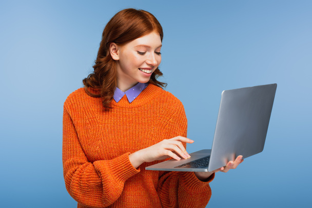 happy redhead student in orange sweater using laptop isolated on blue - Photo, Image
