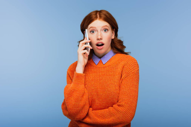 surprised redhead woman in orange sweater talking on smartphone isolated on blue - Foto, imagen