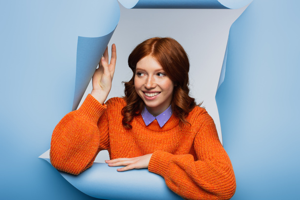 happy young woman in orange sweater looking away on blue ripped background - Фото, зображення