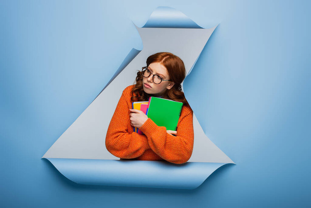 redhead student in orange sweater and glasses holding textbooks on blue ripped paper background  - 写真・画像