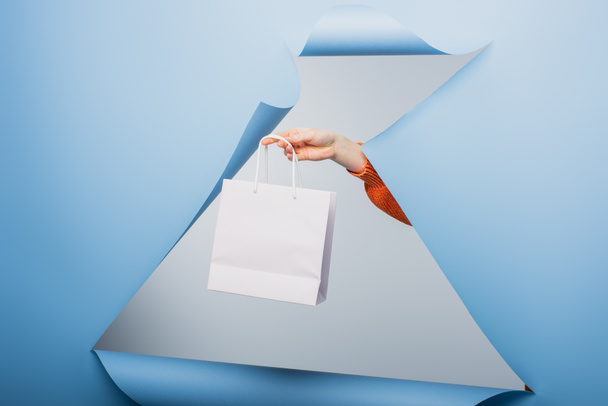 cropped view of woman holding white shopping bag on blue ripped paper background  - Foto, afbeelding