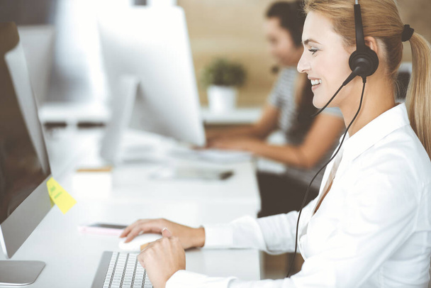 Blonde business woman using headset for communication and consulting people at customer service office. Call center. Group of operators at work - Fotó, kép