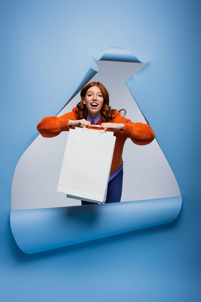 κατάπληκτος νεαρή γυναίκα κρατώντας τσάντα ψώνια σε μπλε άρπαξαν φόντο χαρτί  - Φωτογραφία, εικόνα