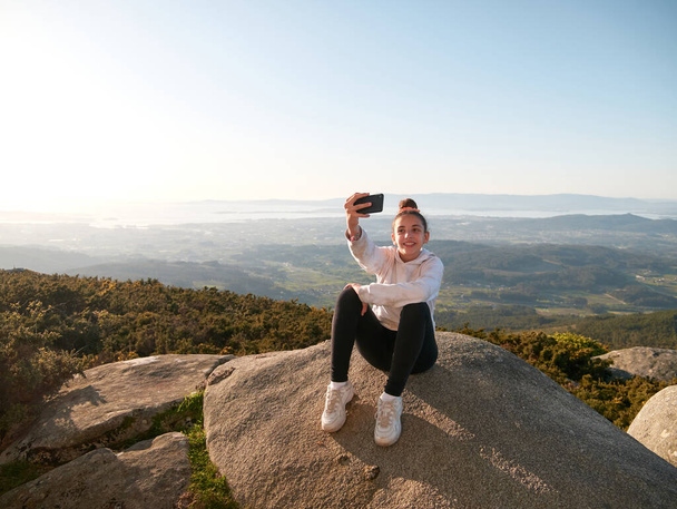 Adolescente tomando una selfie con su teléfono móvil mientras se sienta en una roca en la cima de una colina - Foto, imagen