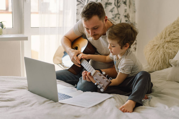 Padre e hijo aprenden a tocar la guitarra acústica en una lección en línea. Tiempo libre para pasar con mi hijo en casa, enseñándole a tocar la guitarra. Feliz día del Padre - Foto, imagen