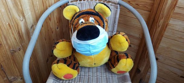 Пандемический ковид-19. мягкий игрушечный тигр в медицинской маске сидит в плетеном кресле - Фото, изображение