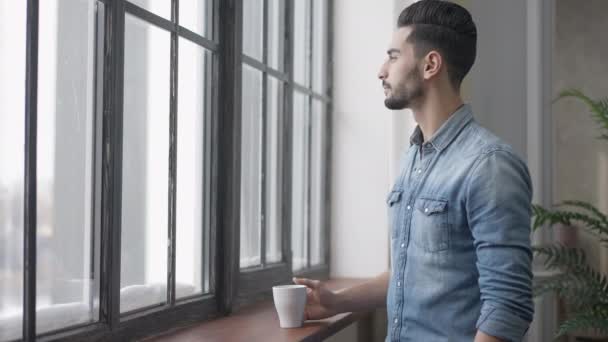 Side view of young handsome Middle Eastern man looking out the window indoors. Portrait of confident fit guy with coffee cup in the morning. Lifestyle concept. - Materiaali, video