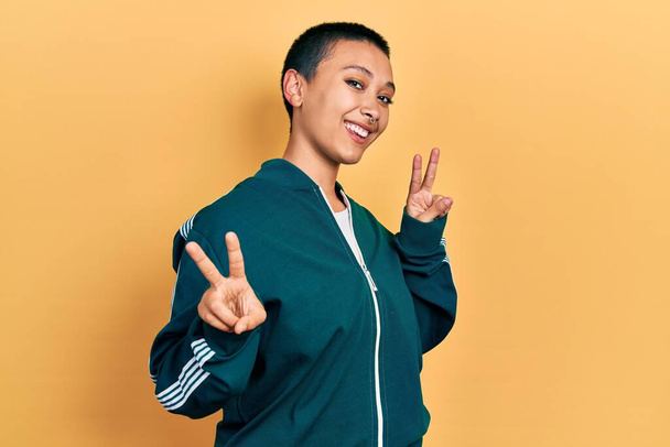 Beautiful hispanic woman with short hair wearing sporty jacket smiling looking to the camera showing fingers doing victory sign. number two.  - Foto, imagen