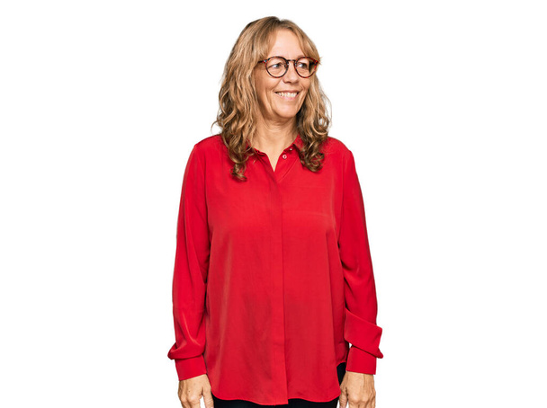 Middle age blonde woman wearing casual shirt over red background looking away to side with smile on face, natural expression. laughing confident.  - Foto, Imagem