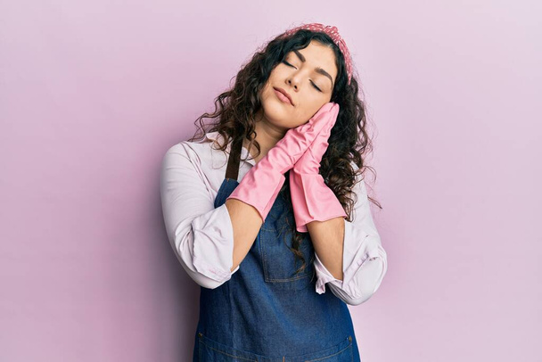 Young brunette woman with curly hair wearing cleaner apron and gloves sleeping tired dreaming and posing with hands together while smiling with closed eyes.  - Photo, image