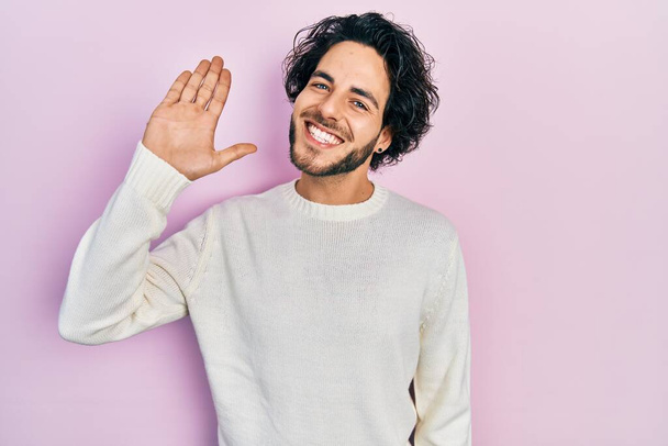 Schöner hispanischer Mann in lässigem weißem Pullover, der fröhlich und lächelnd grüßt, freundliche Willkommensgeste  - Foto, Bild