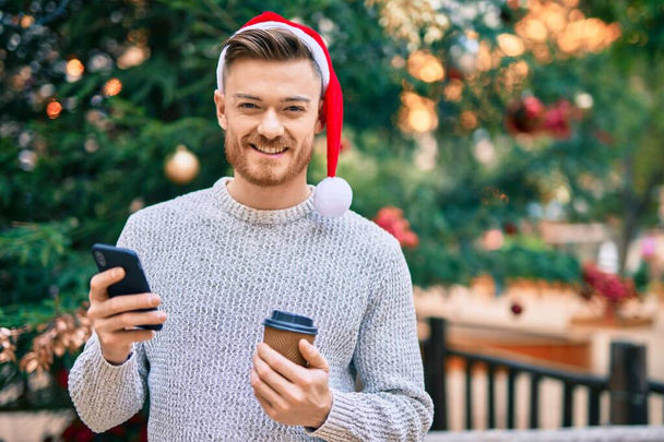 Joven hombre caucásico con sombrero de navidad usando teléfono inteligente y beber café en el parque - Foto, Imagen