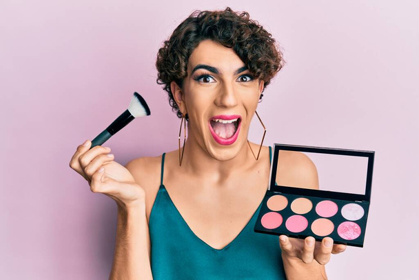 Young man wearing woman make up holding makeup brush and blush celebrating crazy and amazed for success with open eyes screaming excited.  - Photo, Image