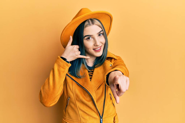 黄色の帽子と革のジャケットを身に着けている若い現代の女の子は、電話のジェスチャーで話しているとあなたを指して笑っています。電話してくれ.  - 写真・画像