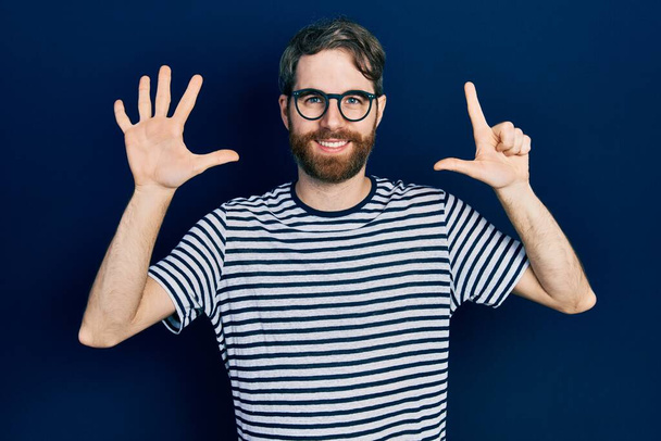 Uomo caucasico con la barba che indossa maglietta a righe e occhiali che mostrano e puntano verso l'alto con le dita numero sette sorridendo fiducioso e felice.  - Foto, immagini
