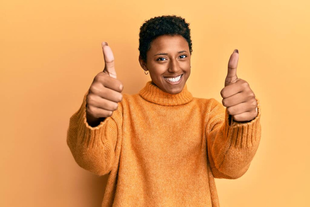 Νεαρή Αφρικάνα Αμερικανίδα που φοράει καθημερινά ρούχα εγκρίνοντας να κάνει θετική χειρονομία με το χέρι, με τους αντίχειρες ψηλά χαμογελώντας και χαρούμενη για την επιτυχία. νικήτρια χειρονομία.  - Φωτογραφία, εικόνα