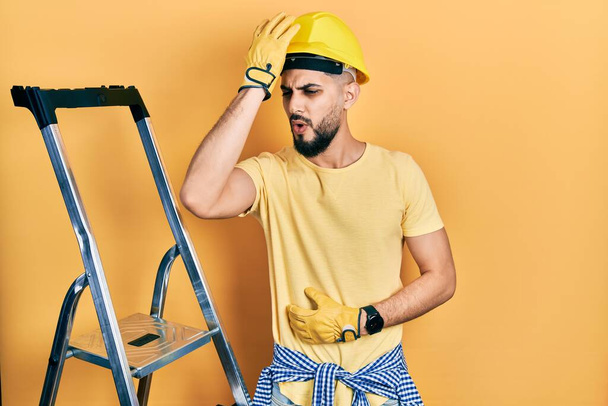 Handsome man with beard by construction stairs wearing hardhat surprised with hand on head for mistake, remember error. forgot, bad memory concept.  - Foto, Imagen