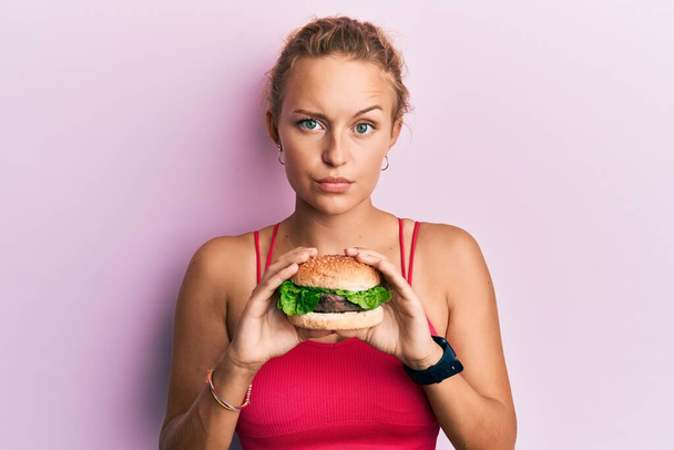 Güzel, beyaz bir kadın lezzetli, klasik bir burger yiyor şüpheci ve gergin, sorun yüzünden surat asıyor. negatif kişi.  - Fotoğraf, Görsel