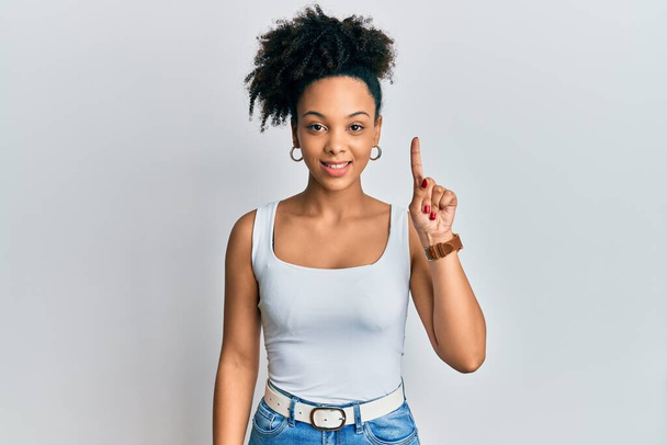 Fiatal afro-amerikai lány hétköznapi stílusban, ujjatlan ingben mosolyog egy ötlettel vagy kérdéssel, ujjal mutogatva boldog arccal, első számú:  - Fotó, kép